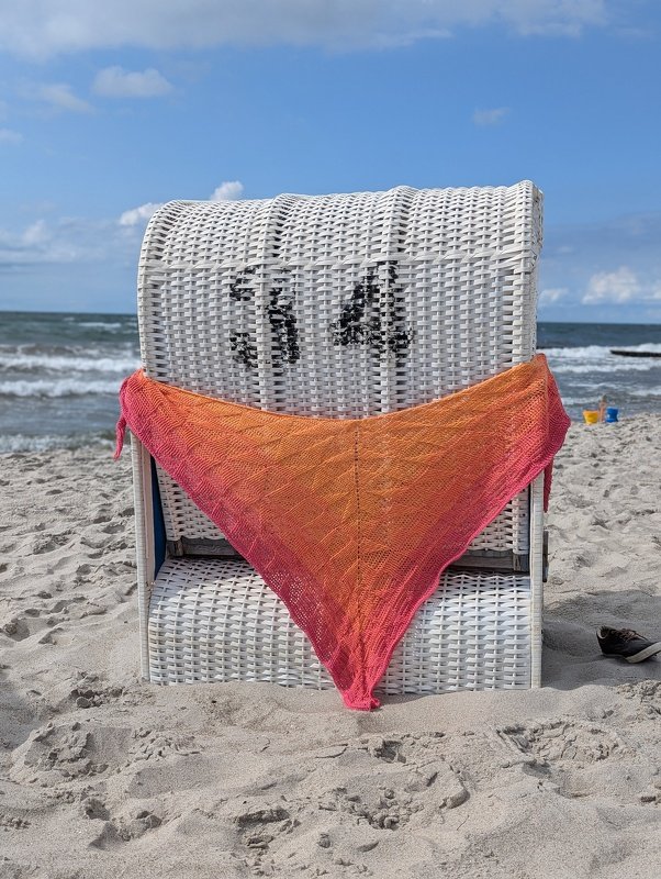 Pink-oranges gehäkeltes Dreieckstuch über einem weißen Strandkorb am Meer.