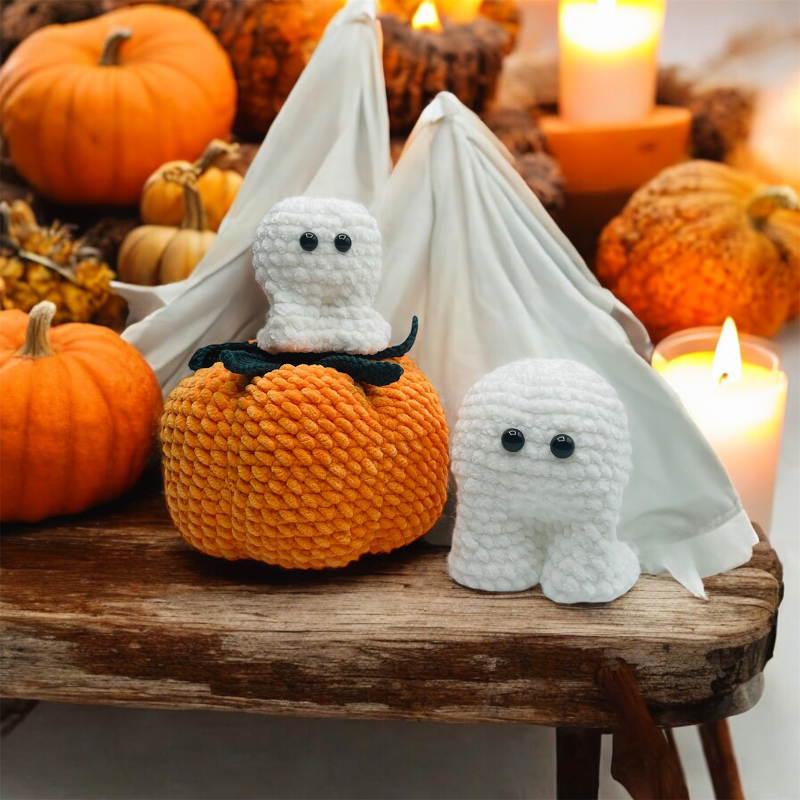 Gehäkelter orangefarbener Kürbis mit zwei weißen Geisterfiguren auf einer Holzbank vor Kerzen