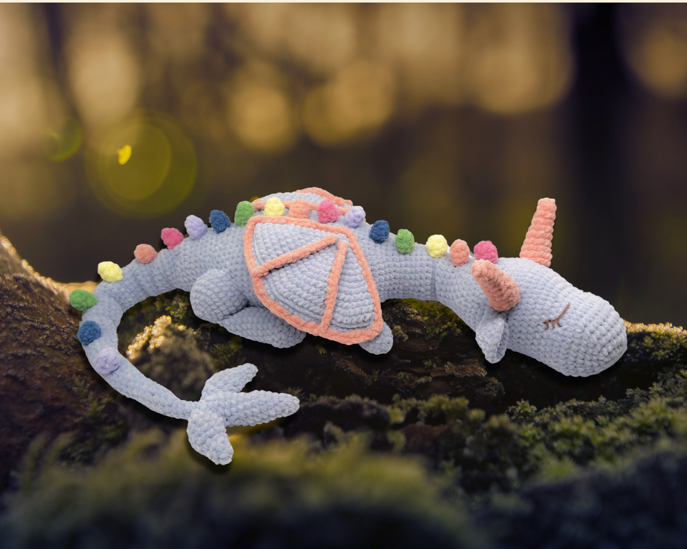 Light blue crocheted dragon amigurumi with pastel bobble spikes and pink horns resting on moss.
