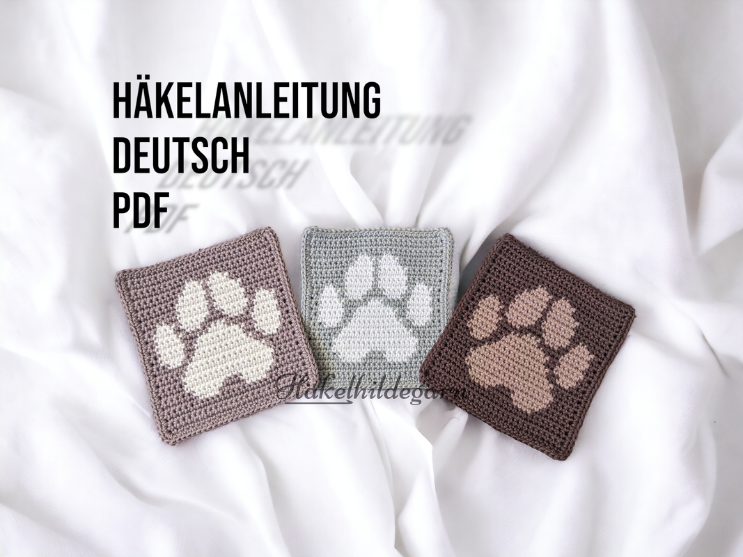 Nahaufnahme von drei gehäkelten Quadraten mit Pfotenmotiv auf weißem Laken.