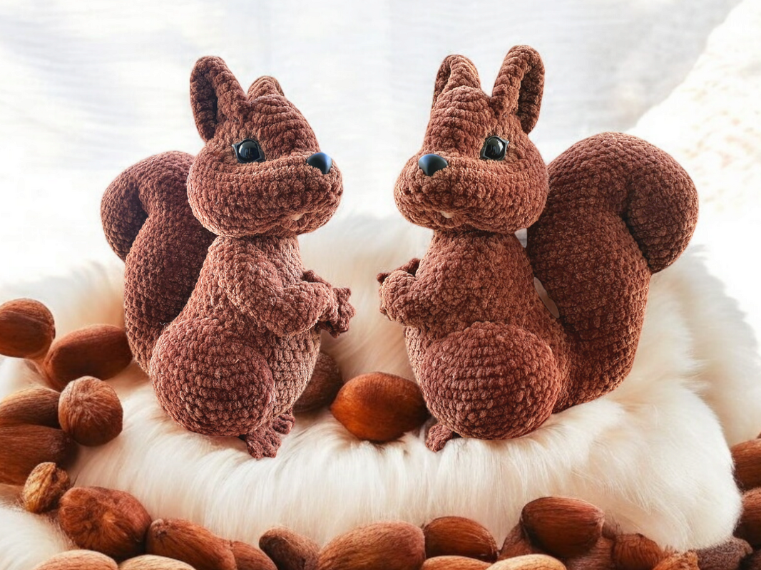 Zwei braune gehäkelte Eichhörnchen-Amigurumi auf weißem Fell, Nüsse liegen rundherum