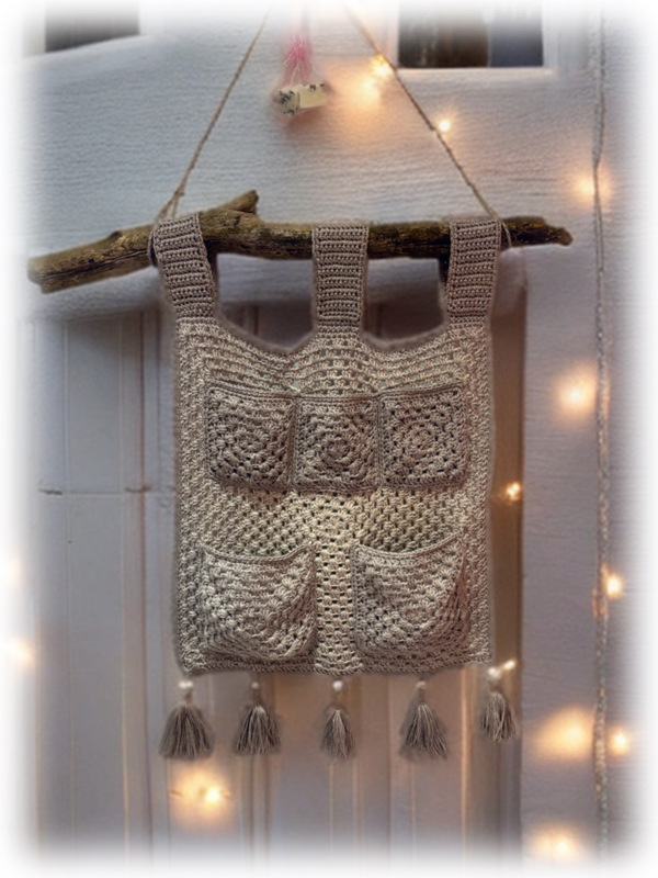 Gehäkelter Boho-Hängeorganizer mit Taschen und Quasten an einem Holzast, im Hintergrund Lichterketten.