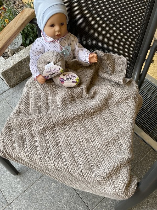 Babypuppe mit beigefarbener gestrickter Babydecke mit diagonalem Rippenmuster und Garnknäueln