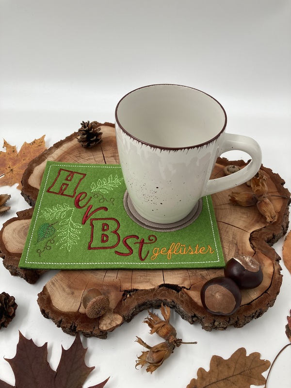 Grüner rechteckiger ITH-Tassenteppich mit gestickten Blättern und Schriftzug 'Herbstgeflüster' neben Keramiktasse, Kastanien und Eicheln