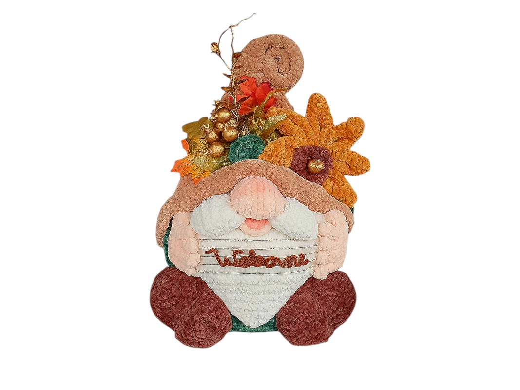 Crocheted gnome wearing a brown hat and holding a welcome sign, decorated with orange leaves, gold berries, and an orange textured flower.