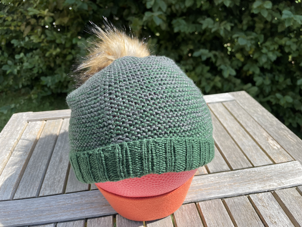 Dunkelgrüne gestrickte Mütze mit braunem Fellbommel auf einem Schaummodel, auf einem hölzernen Tisch