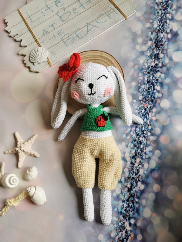 Hand-crocheted white rabbit with a red head accessory, green top with ladybug applique, and beige shorts displayed with seashells and beach decorations.
