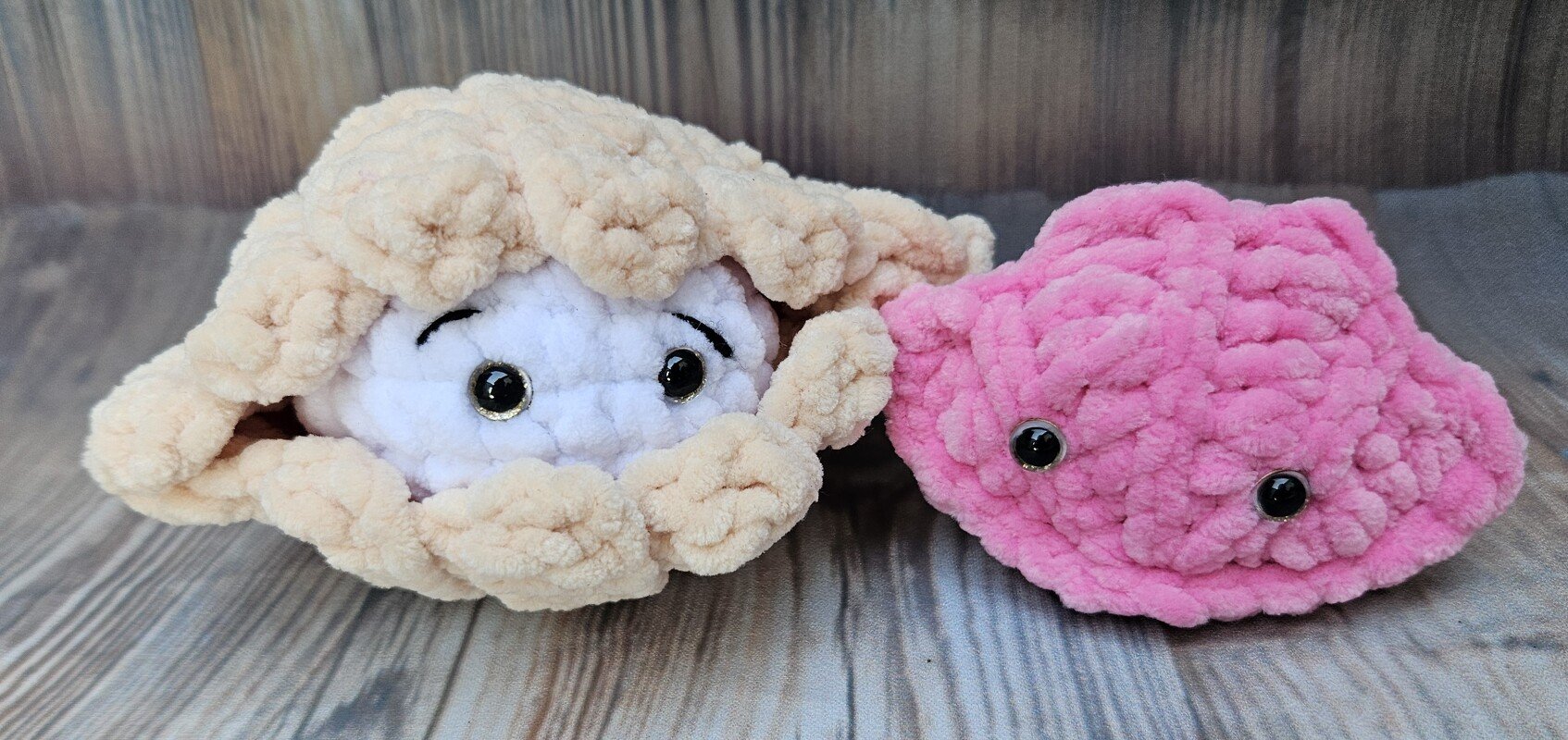 Zwei gehäkelte Muschelamigurumis auf Holzuntergrund: beige Muschel mit weißem Innenkörper und glänzenden Augen, daneben rosa Muschel
