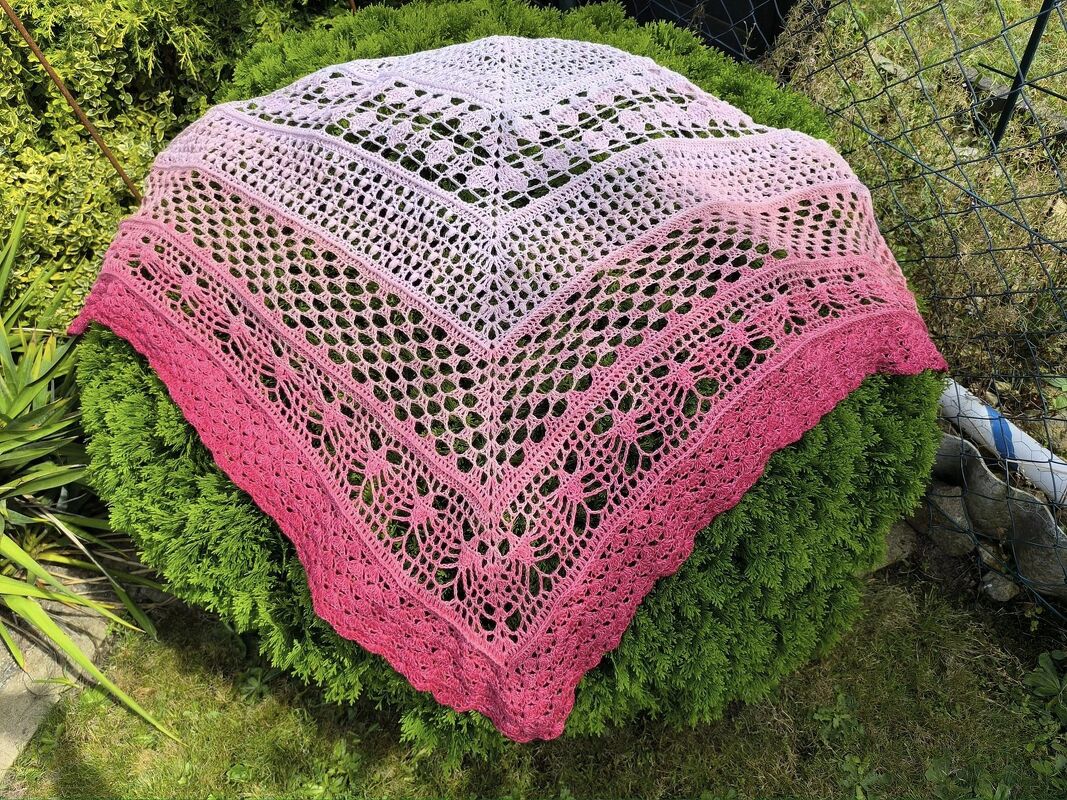 Gehäkeltes Dreieckstuch in Rosa‑Fuchsia mit durchbrochenem Muster auf grünem Strauch