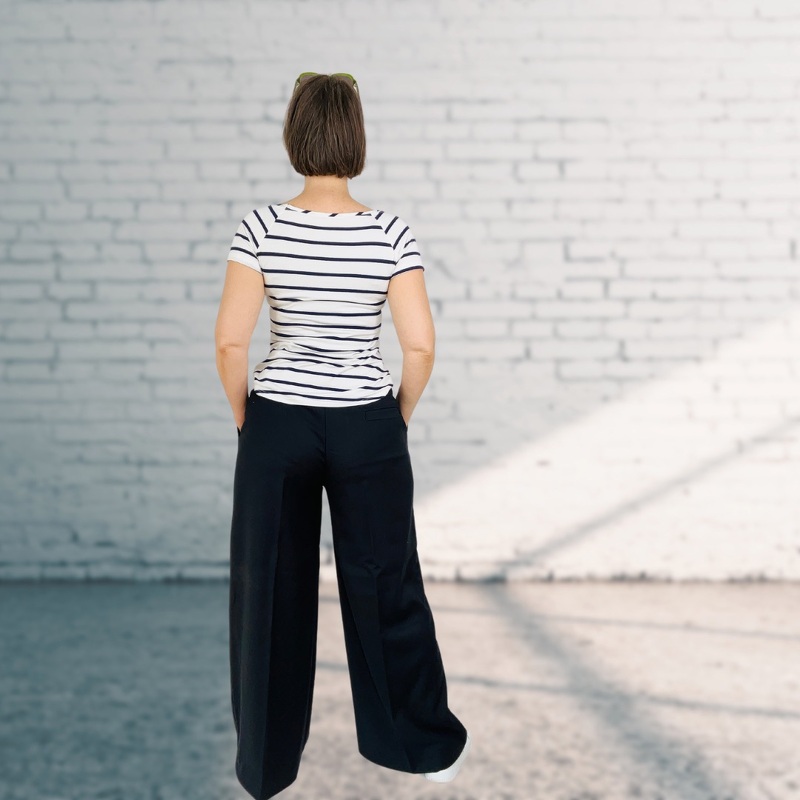 Rückenansicht: weiß-blau gestreiftes Kurzarmshirt und dunkle weite Hose, Hände in den Taschen