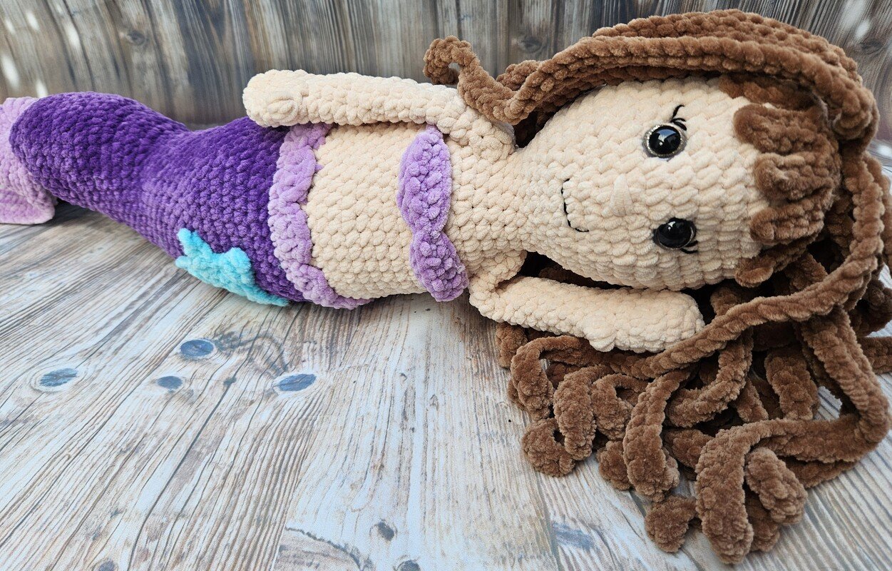 Gehäkelte Meerjungfrau-Amigurumi mit lila Schwanz und braunen Locken auf Holzuntergrund