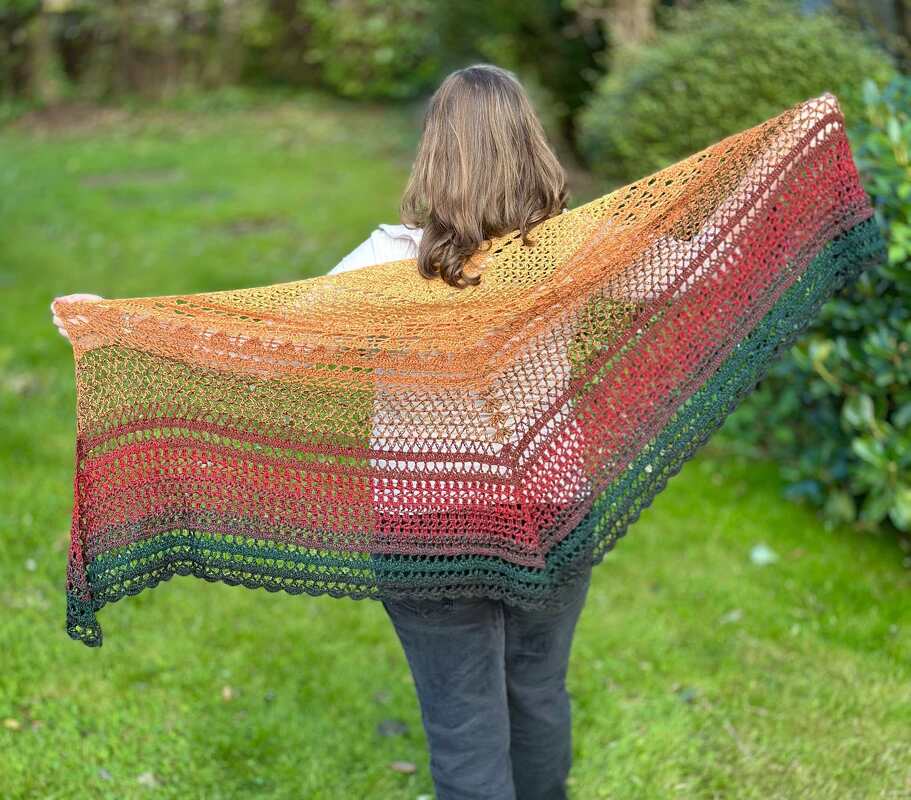 Rückenansicht einer Person mit großem, mehrfarbigem gehäkeltem Dreieckstuch in herbstlichen Tönen im Garten