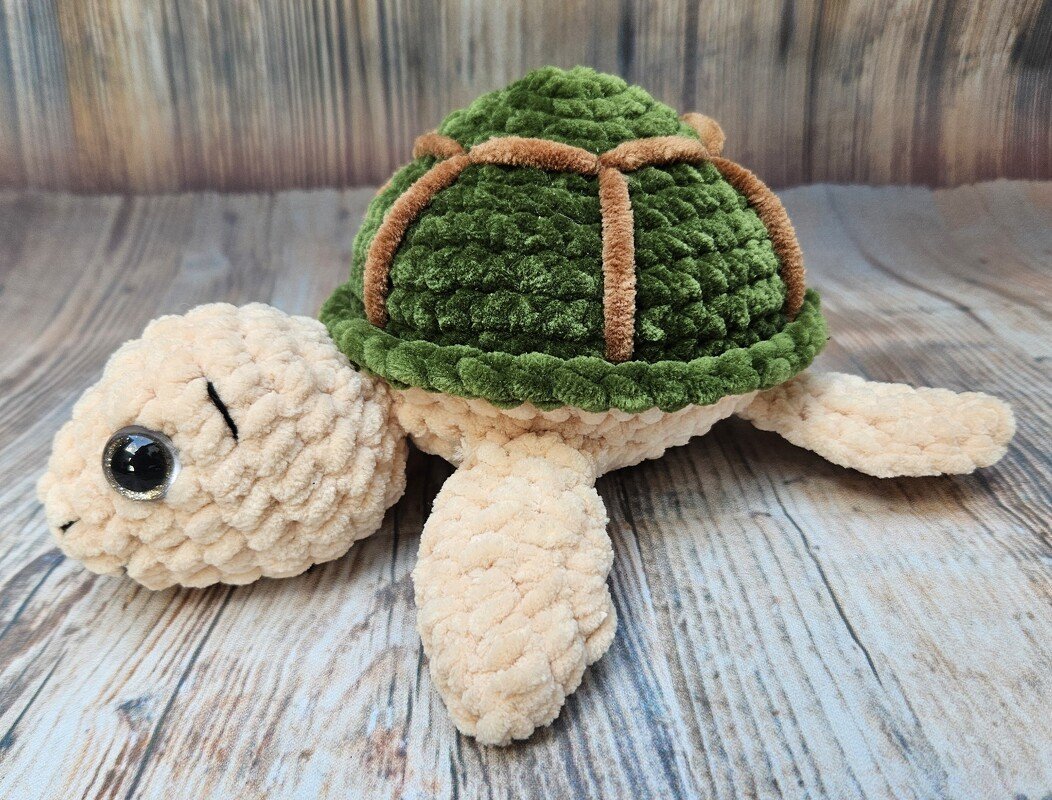 Gehäkelte Amigurumi‑Schildkröte mit grünem Panzer und beigem Körper auf Holzboden.