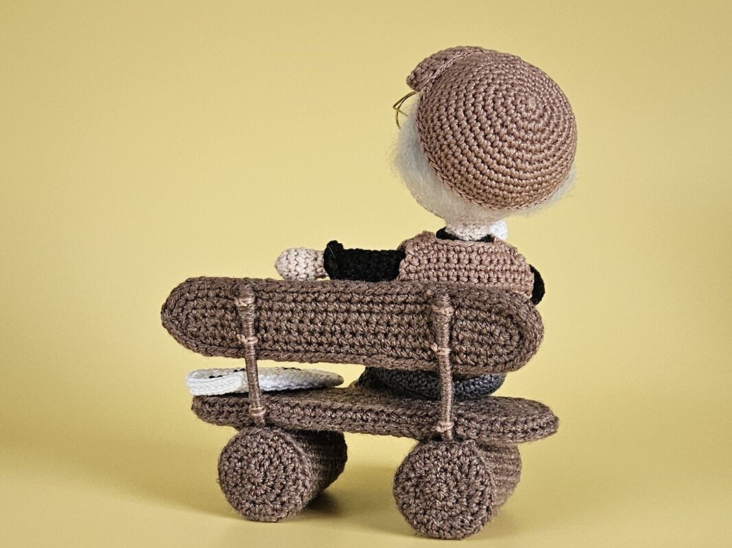 Gehäkelte Amigurumi-Puppe sitzt von hinten auf einer braunen Bank mit Rädern vor gelbem Hintergrund.