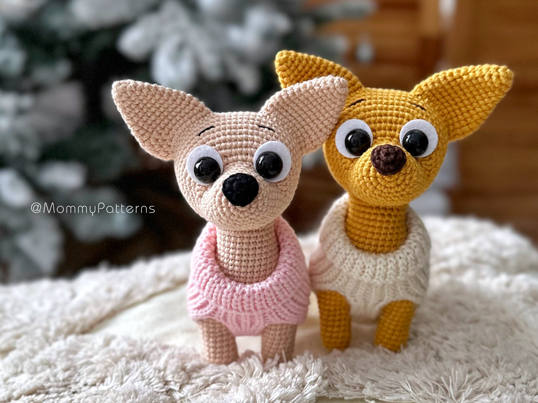 Zwei gehäkelte Amigurumi‑Hunde, einer rosa mit rosa Pullover, einer senfgelb mit cremefarbenem Pullover, auf flauschigem Untergrund
