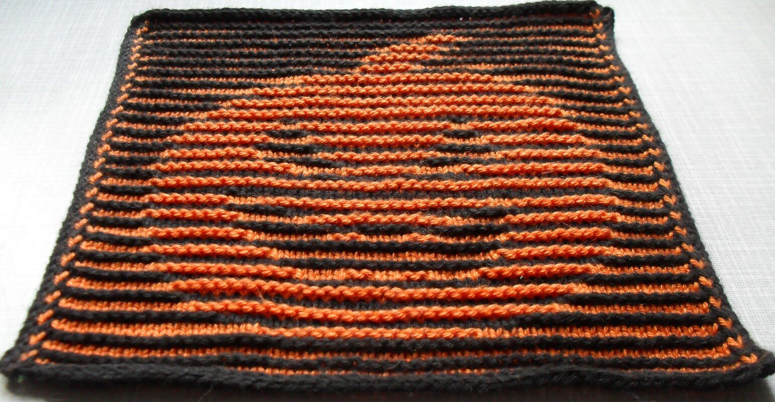 Gestricktes quadratisches Dekostück in Schwarz und Orange mit Kürbismotiv auf hellem Untergrund
