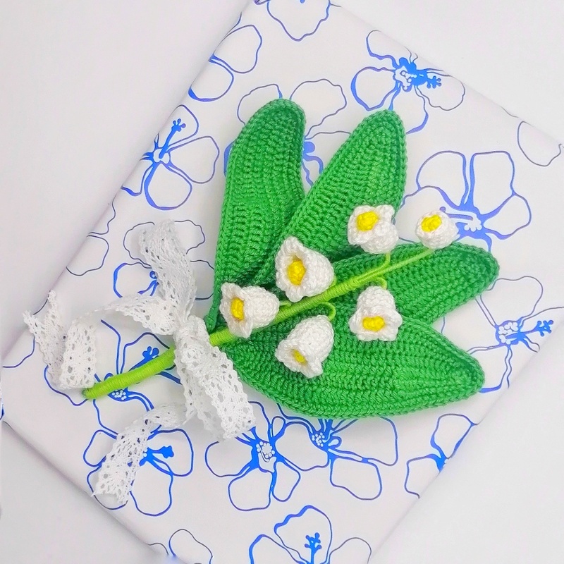 Nahaufnahme einer gehäkelten Maiglöckchen-Applikation mit grünen Blättern, weißen Glockenblüten und weißem Spitzenband auf gemustertem Papier.