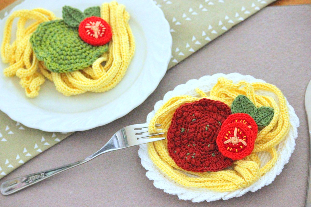 Zwei weiße Teller mit gehäkelten gelben Spaghetti, roten Tomatenscheiben und grünen Blättern; eine Gabel liegt auf einem Teller