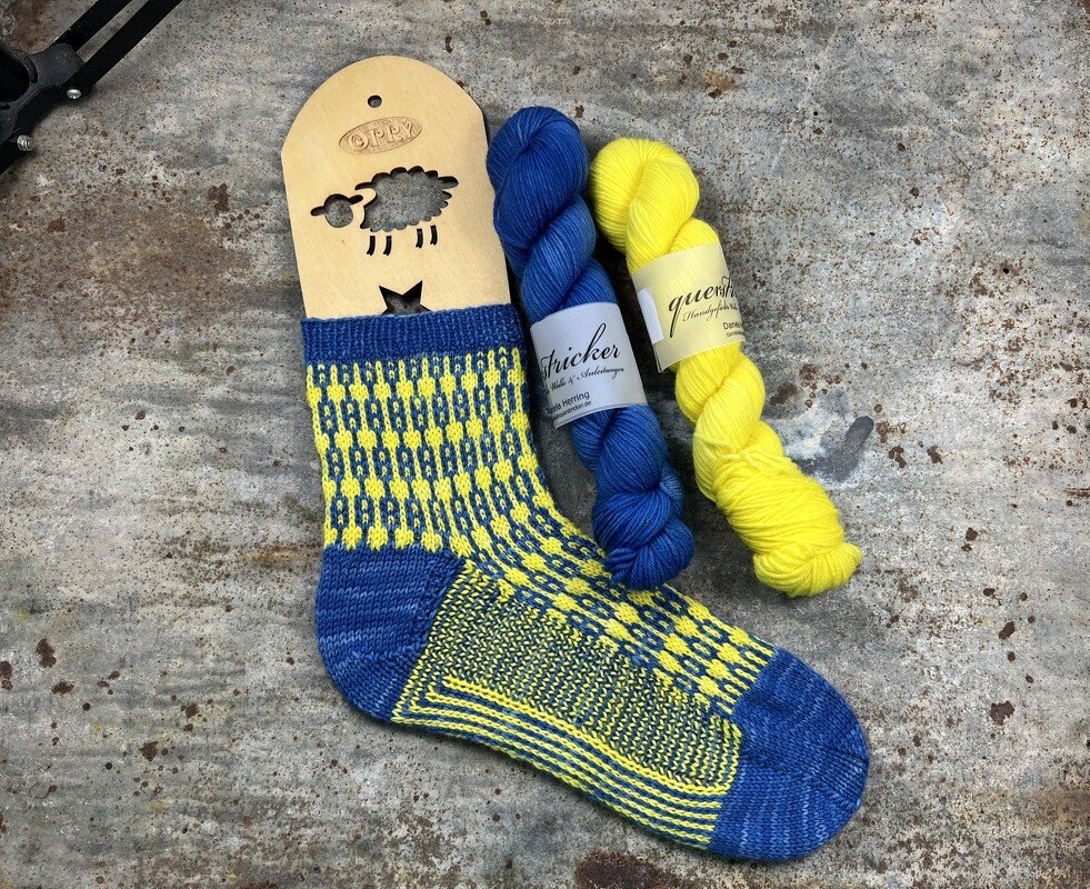 Blau-gelbe gestrickte Damensocke auf Holz-Sockenblocker, daneben zwei Garnstränge in Blau und Gelb