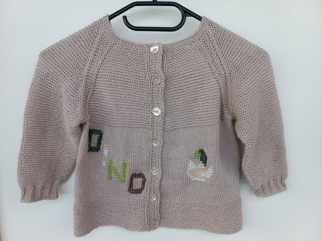 Beige gestrickte Babyjacke mit DINO-Stickerei und Dinosaurier-Ei, an einem Kleiderbügel