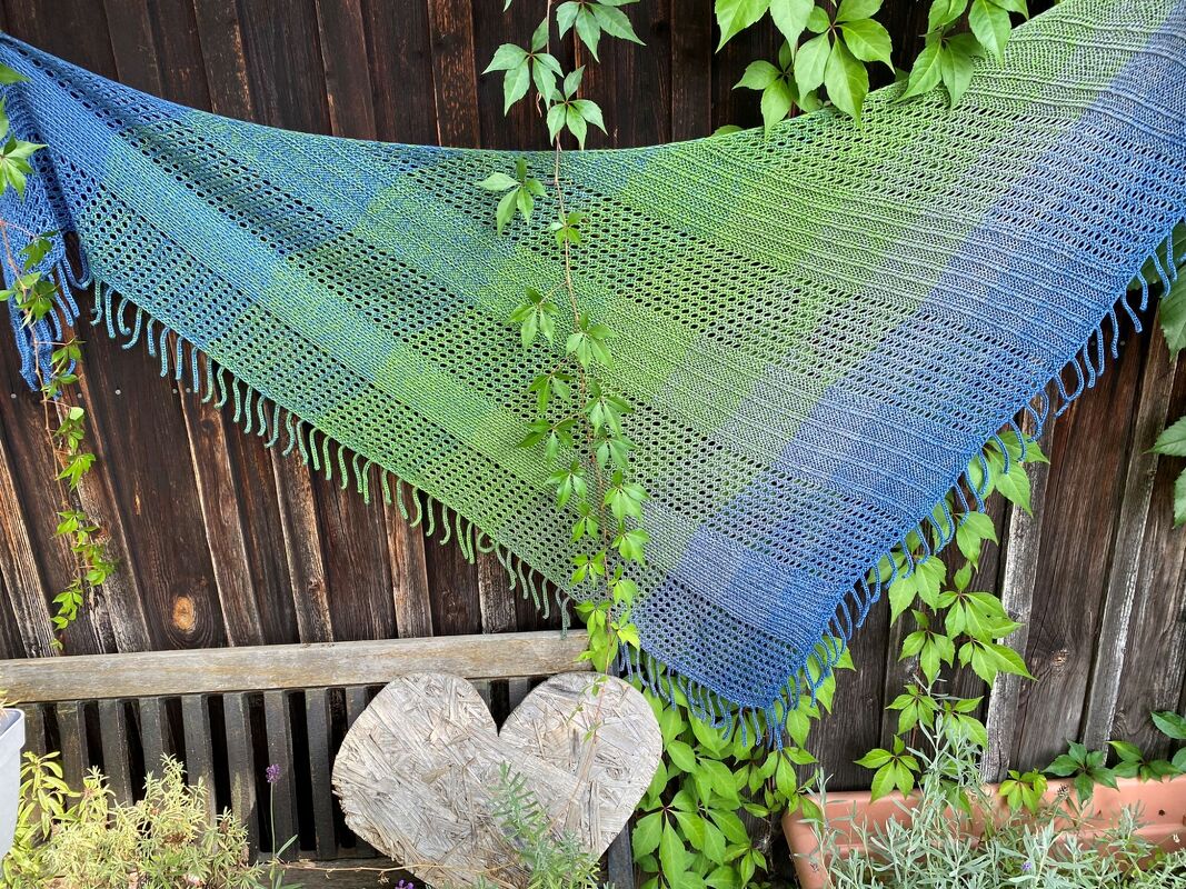 Blau-grünes gestricktes Dreieckstuch mit Lochmuster und Fransen, an einem Holzzaun aufgehängt.
