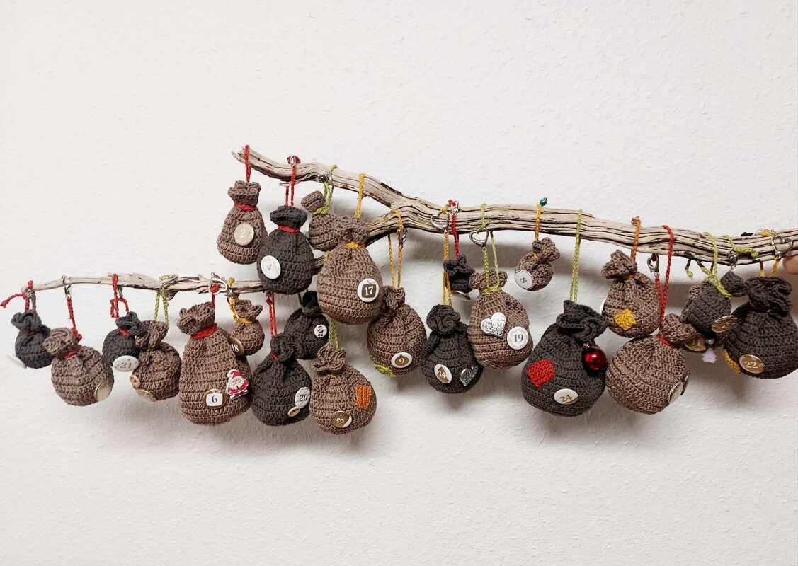 Gehäkelter Adventskalender mit 24 nummerierten Säckchen an Ästen vor weißer Wand
