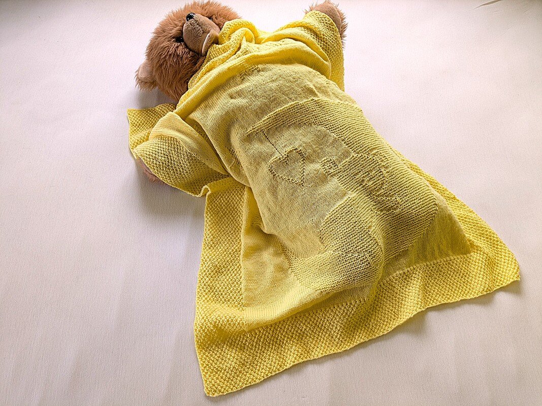 Teddy bear partially covered by a bright yellow knitted baby blanket with heart and bottle motifs.