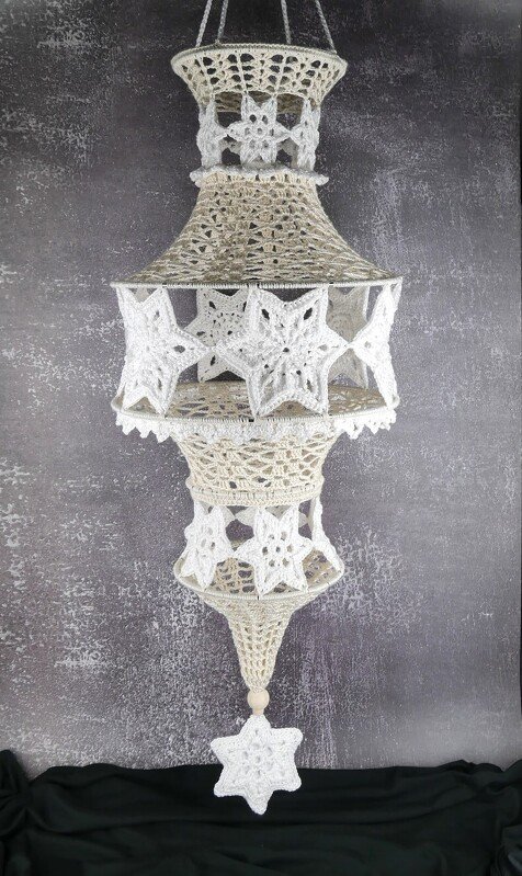 Mehrstufiger gehäkelter Lampion in Beige und Weiß mit Sternmotiven, hängend vor dunklem, strukturiertem Hintergrund.