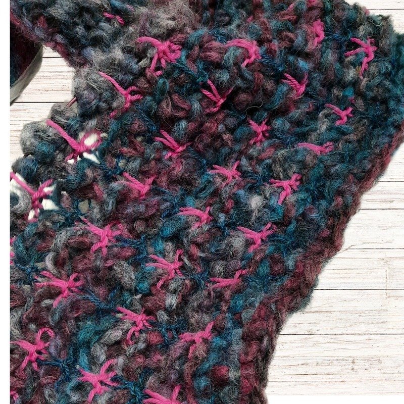 Grob gestrickter Mehrfarbenschal in Grau, Türkis und Beere mit pinken Kreuzstichen auf hellem Holz