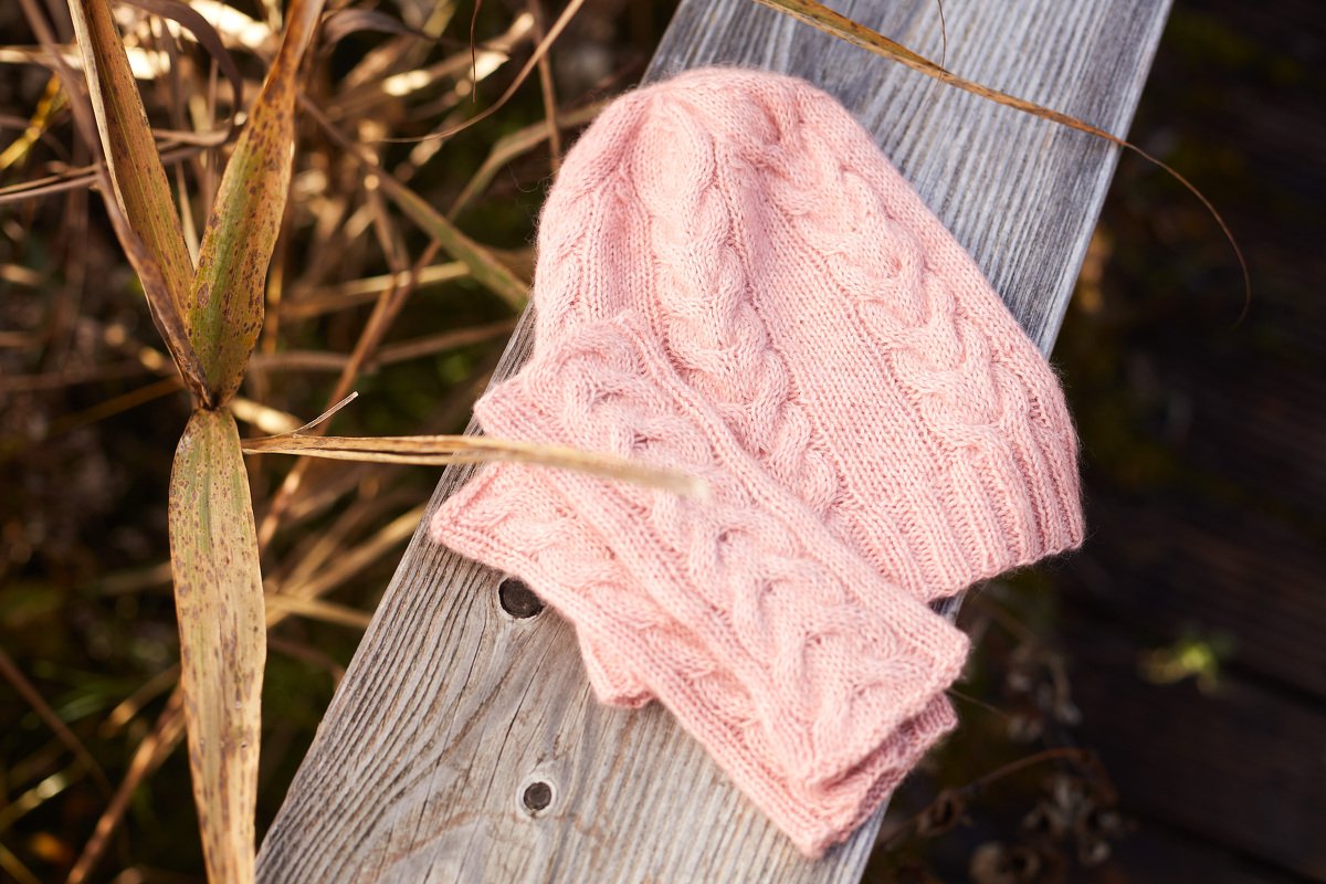 Rosa gestrickte Mütze und passender Loop mit Zopfmuster auf einer Holzbank