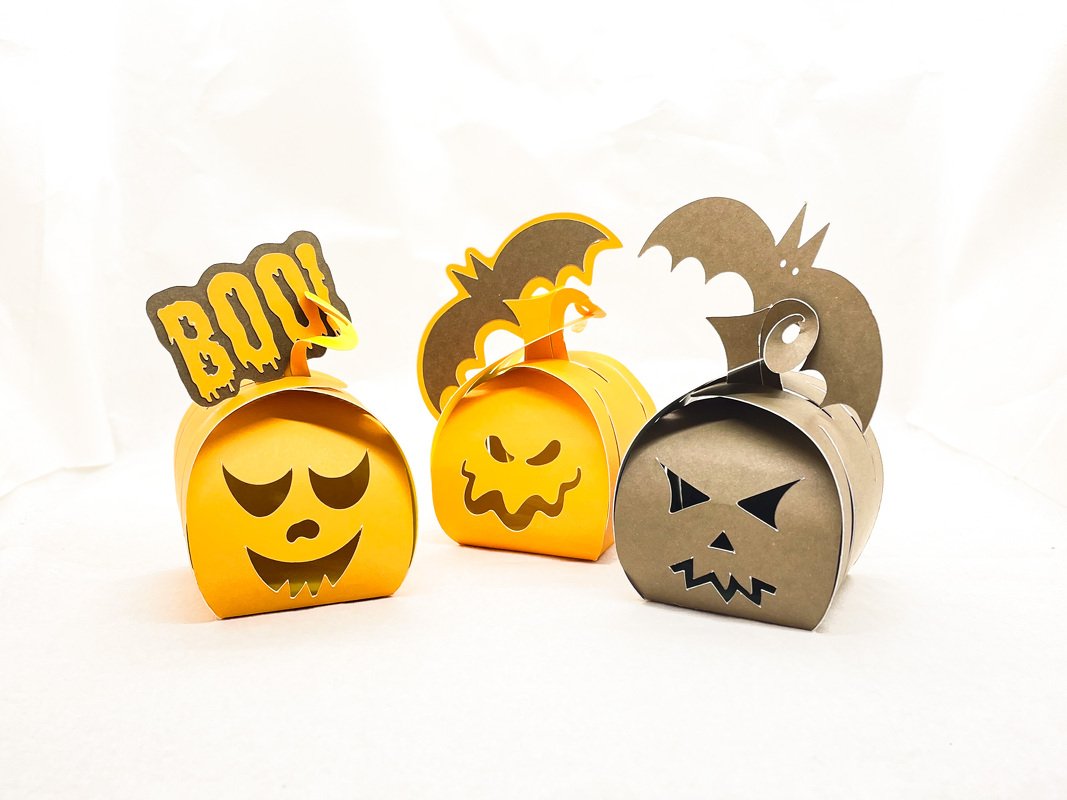 Drei gebastelte Halloween-Kürbis-Füllboxen (zwei orange, eine grau) mit ausgestanzten Gesichtern, Fledermaus- und 'BOO!'-Elementen.