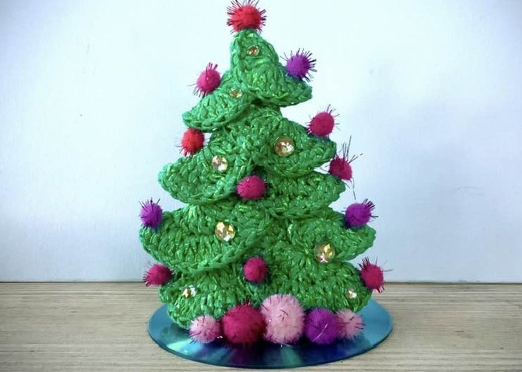 Gehäkelter Mini-Weihnachtsbaum mit bunten Bommeln und goldenen Pailletten auf runder Spiegelunterlage