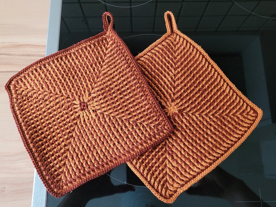 Zwei quadratische gehäkelte Topflappen in Orange und Braun mit diagonaler Rippenstruktur und Aufhängeschlaufen