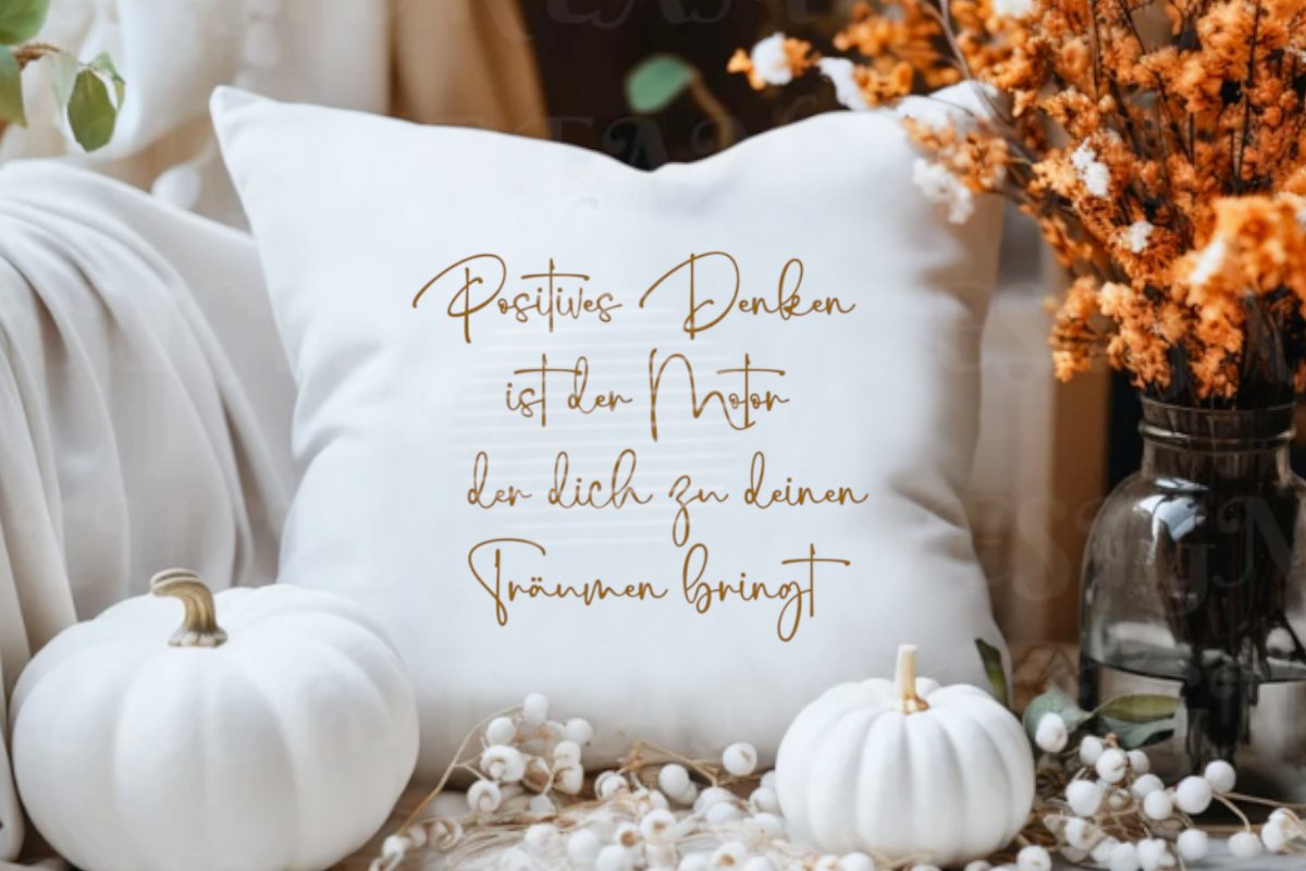 Nahaufnahme eines weißen Kissens mit dem Zitat ‚Positives Denken ist der Motor der dich zu deinen Träumen bringt‘, umgeben von weißen Kürbissen und herbstlicher Deko.