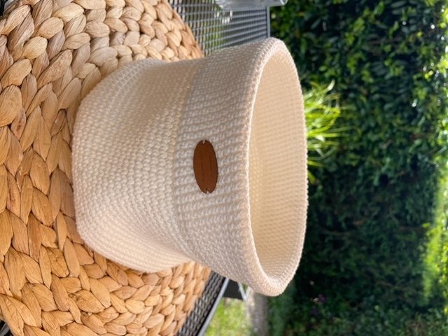 Cremefarbener gehäkelter Korb mit braunem Anhänger auf geflochtener Matte vor grünem Hintergrund