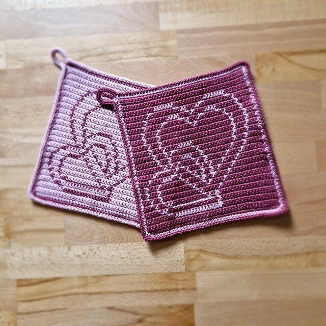 Zwei gehäkelte quadratische Topflappen in Rosa und Bordeaux mit Herz- und Friedensmotiv auf Holz