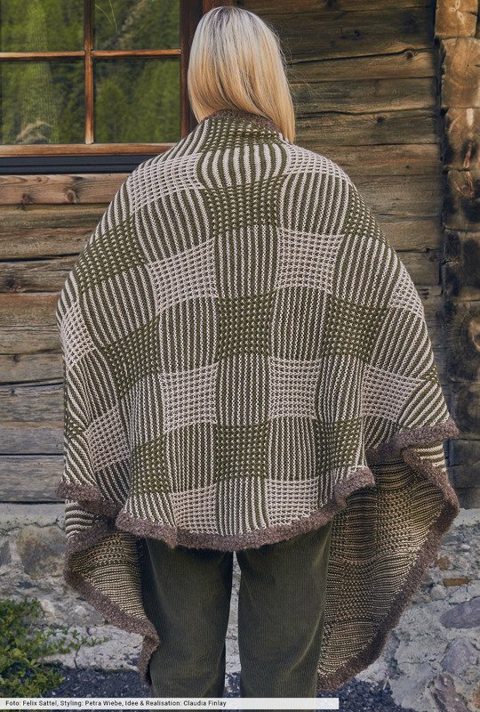 Rückenansicht einer Person mit hellem Haar, die einen gestrickten Poncho mit grün-beigem Schachbrettmuster und dunkelbrauner Kante trägt.