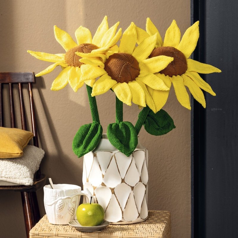 Drei gestrickte Sonnenblumen mit gelben Blütenblättern und braunen Kernen in einer weißen, strukturierten Vase, daneben eine Tasse und ein grüner Apfel auf einem geflochtenen Beistelltisch.