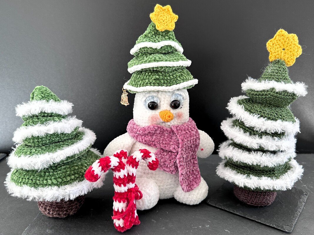 Gehäkelter Schneemann mit grüner Tannenmütze, rosa Schal und gehäkelter Zuckerstange, flankiert von zwei kleinen gehäkelten Weihnachtsbäumen mit weißen Borten.