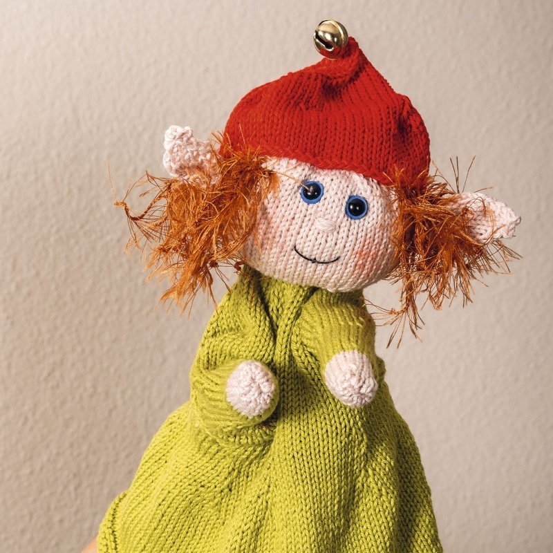 Gestrickte Wichtel-Figur mit grünem Kleid, roter Zipfelmütze mit Glöckchen, roten Fransenhaaren und blauen Knopfaugen.