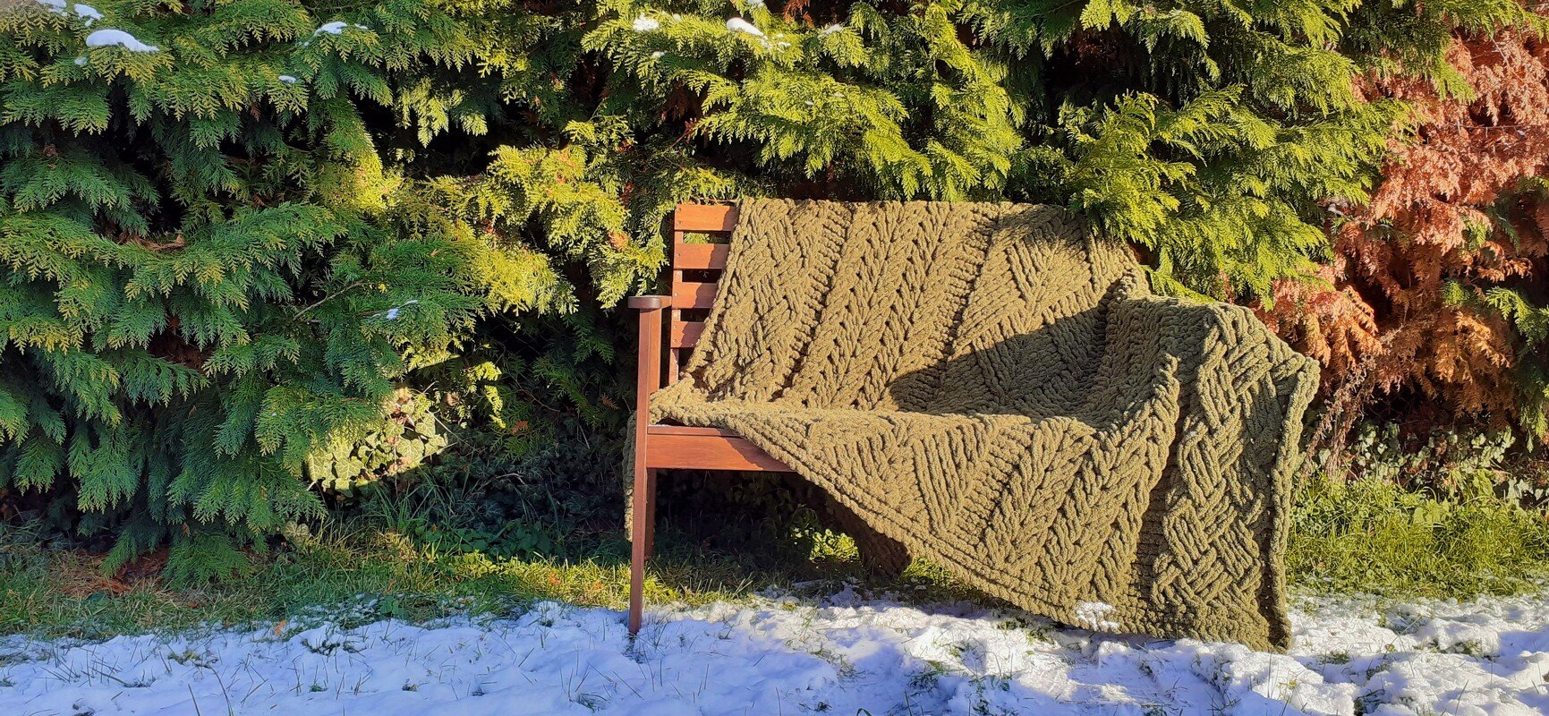 Grob gehäkelte Zopfdecke mit sichtbaren Maschenreihen auf einer Holzbank vor grüner Hecke und leichtem Schnee
