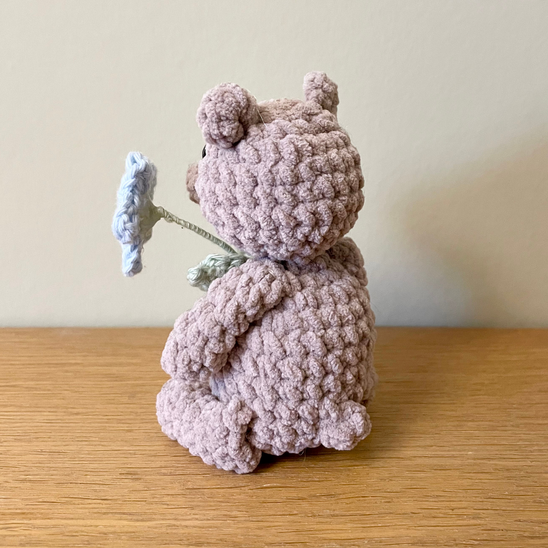 Rückenansicht: sitzender gehäkelter Teddy in zartrosa mit Blume
