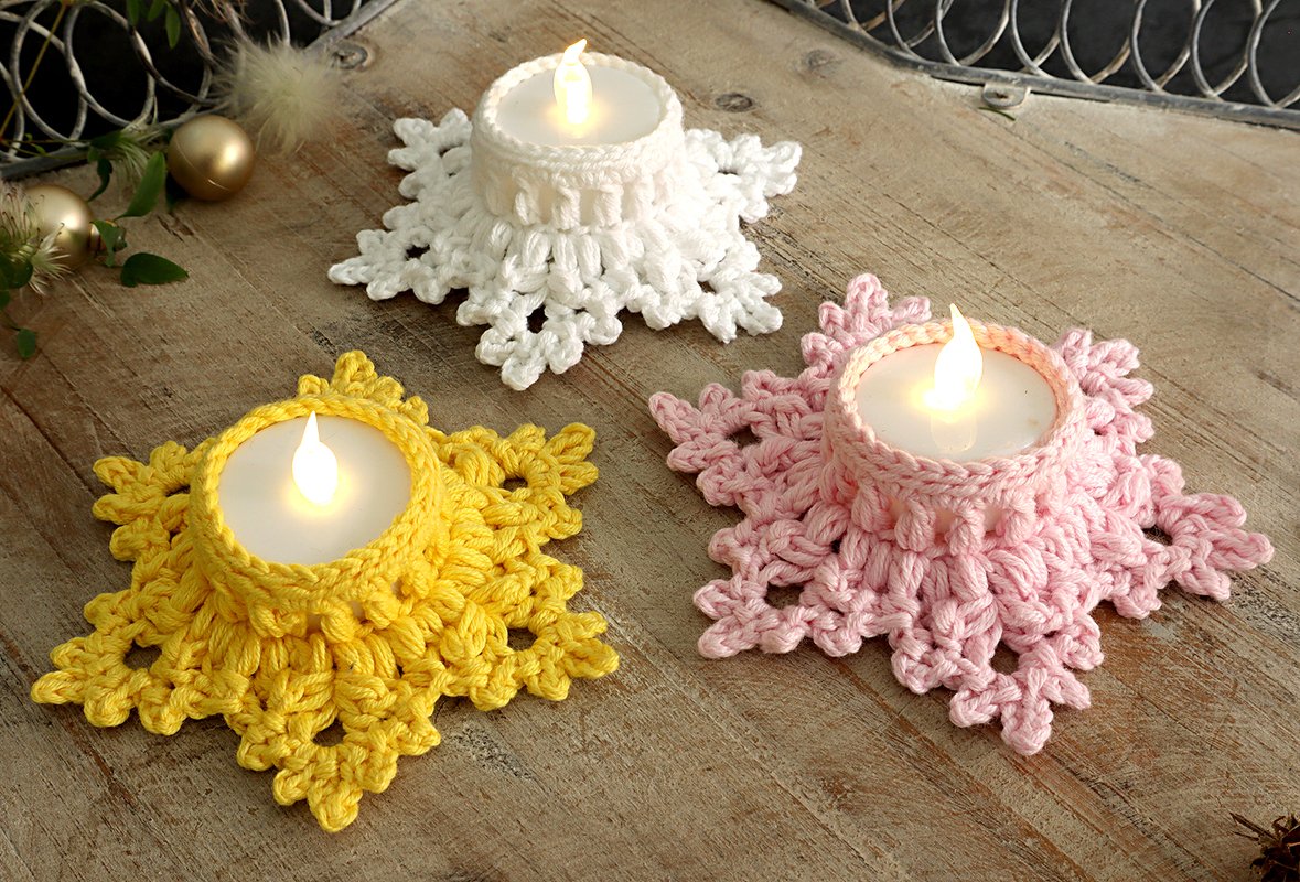 Drei gehäkelte Schneeflocken-Teelichthalter (weiß, gelb, rosa) mit LED-Teelichtern auf Holztisch.