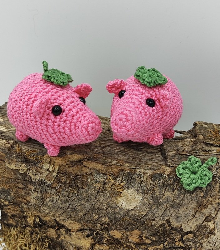 Zwei rosa gehäkelte Amigurumi-Schweinchen mit grünen Blättern auf einem Holzstamm, daneben ein grünes gehäkeltes Kleeblatt