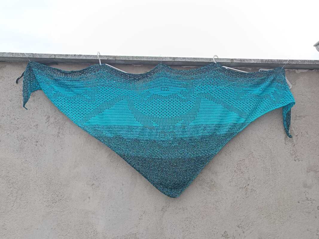 Gehäkeltes Dreieckstuch in Türkis und Grau mit zentralem Herz-Lochmuster, an Wand aufgehängt