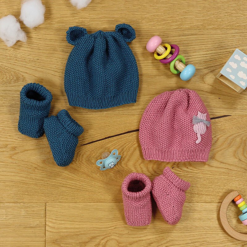 Zwei gestrickte Babysets auf Holzuntergrund: blaues Mützchen mit Öhrchen und Söckchen, rosa Mütze mit Katzenapplikation und passende Söckchen, umgeben von Babyspielzeug