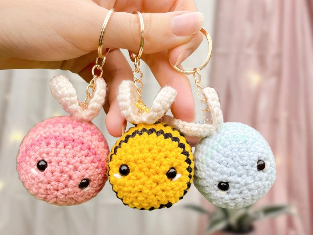 Hand hält drei kleine, schnell gehäkelte runde Amigurumi-Anhänger in Rosa, Gelb und Hellblau mit kleinen Ohren an goldenen Ringen.