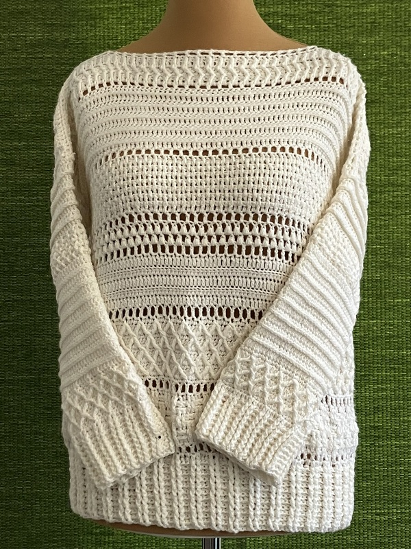 Cremefarbener gehäkelter Damenpullover mit langen Ärmeln auf einer Schneiderpuppe vor grünem Hintergrund.