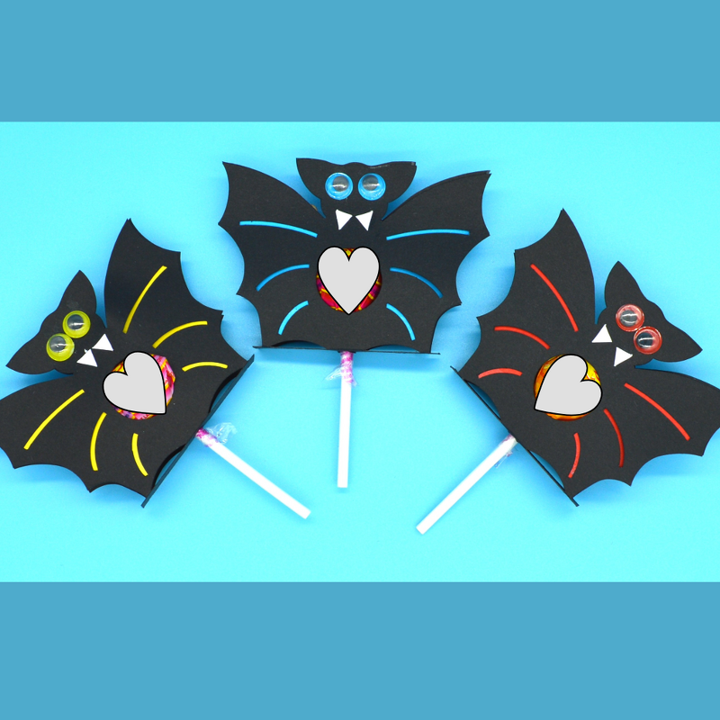 Drei schwarze Papierfledermäuse mit Wackelaugen, die jeweils einen Lutscher in einer herzförmigen Aussparung halten, auf blauem Hintergrund.