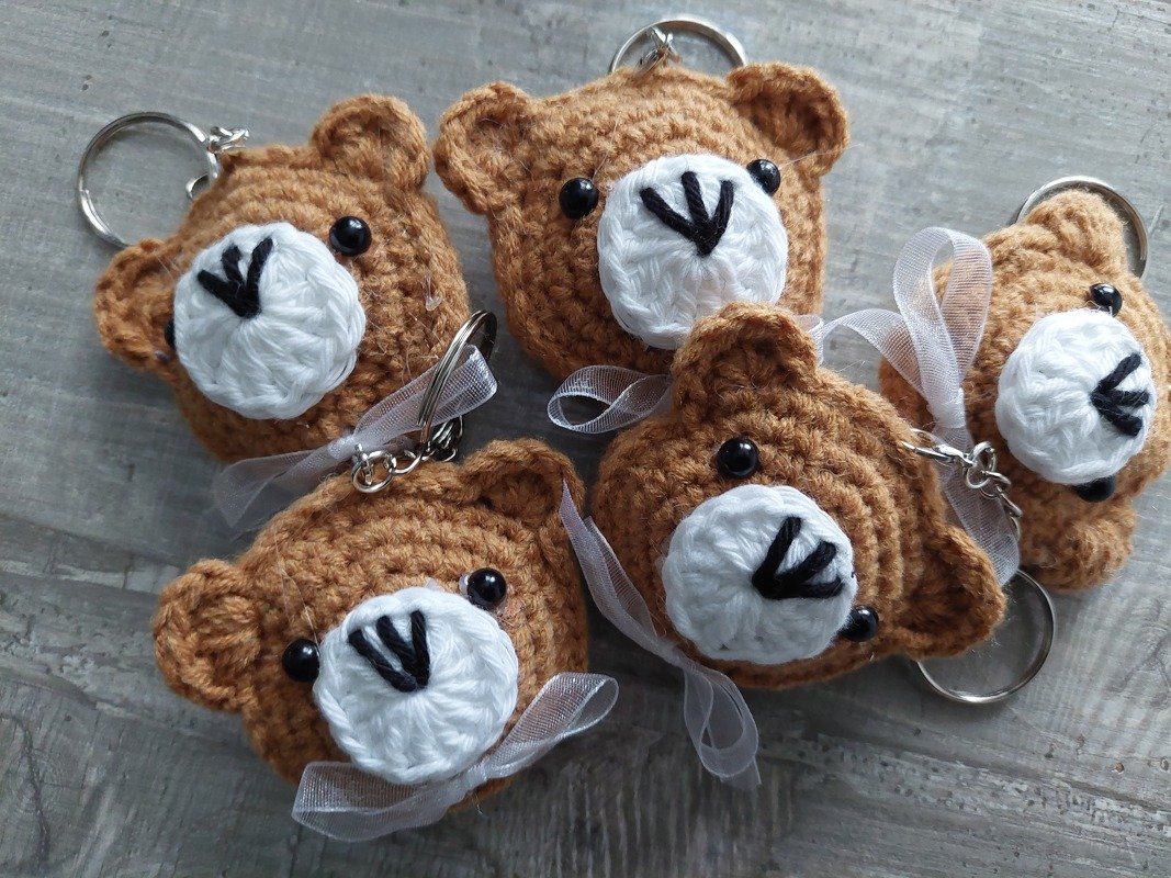 Fünf gehäkelte braune Teddybär-Schlüsselanhänger mit weißen Schnauzen und Schleifen auf grauem Holz.